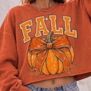 May include: A burnt orange cropped sweatshirt featuring the word FALL in large, distressed block letters above a graphic of a pumpkin tied with a prominent bow. The design uses a textured, weathered aesthetic with orange and white tones.