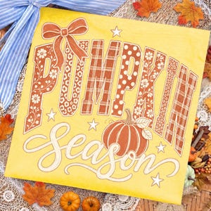 May include: A yellow square sign featuring the text PUMPKIN SEASON in large block letters with floral, plaid, and polka dot patterns. A decorative bow sits above the word PUMPKIN, and a pumpkin illustration is placed next to the word SEASON.