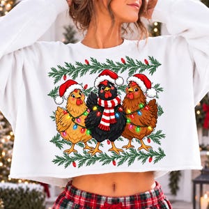 Puede incluir: Una sudadera blanca de manga larga con un gr&aacute;fico de tres pollos con gorros de Pap&aacute; Noel rojos y blancos. Los pollos est&aacute;n envueltos en guirnaldas de luces navide&ntilde;as de colores y el pollo central lleva una bufanda a rayas rojas y blancas.