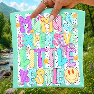 May include: A light green fabric item featuring the text MAMA'S EXPENSIVE LITTLE BESTIE in colorful, patterned block letters. The design includes black and white vertical stripes, small yellow flowers, pink bows, and a yellow smiley face.