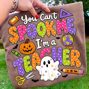 May include: A brown t-shirt featuring a colorful graphic with the text "You Can't SPOOK ME I'm a TEACHER". The design includes a jack-o'-lantern, a ghost with flower accents, a globe, scissors, a pencil, a ruler, an eraser, and small bats.