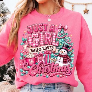 May include: A bright pink sweatshirt featuring a graphic print with the text "JUST A GIRL WHO LOVES Christmas". The design includes a decorated Christmas tree, a snowman wearing a plaid scarf, wrapped gift boxes, and a mug with a snowflake.