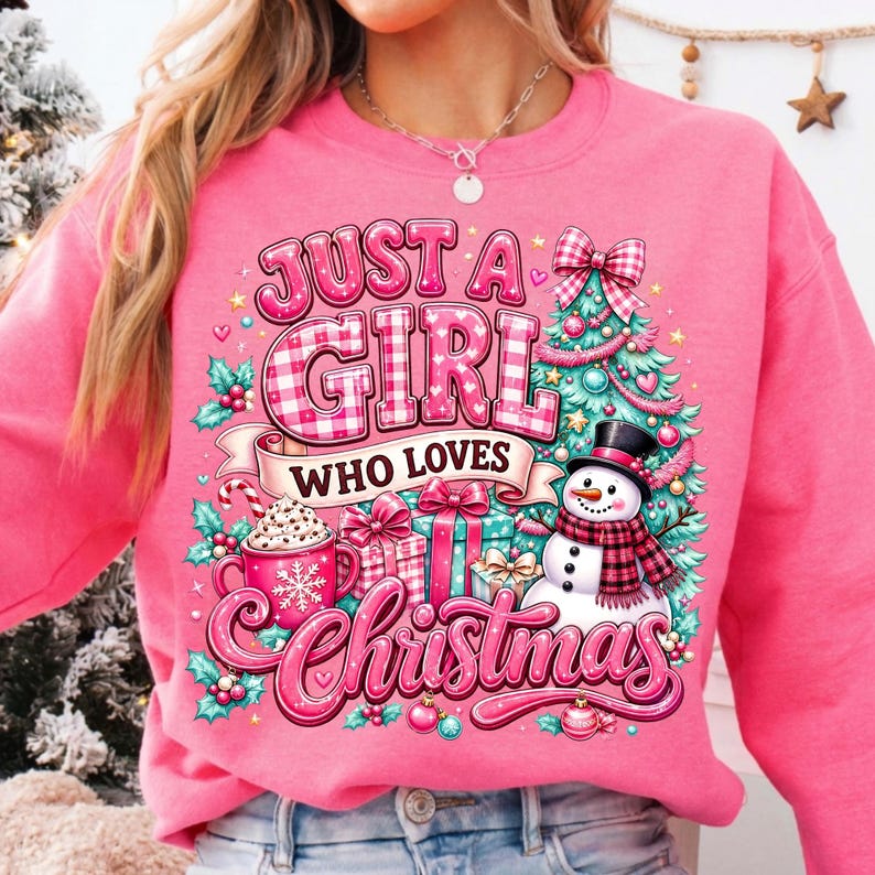 May include: A bright pink sweatshirt featuring a graphic print with the text "JUST A GIRL WHO LOVES Christmas". The design includes a decorated Christmas tree, a snowman wearing a plaid scarf, wrapped gift boxes, and a mug with a snowflake.