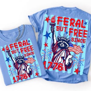 Może przedstawiać: Jasnoniebieski t-shirt z grafiką szopa pracza przebranego za Statuę Wolności, trzymającego pochodnię i książkę. Napis głosi FERAL BUT FREE SINCE 1776. Wz&oacute;r zawiera czerwone i niebieskie gwiazdy, fajerwerki oraz amerykańską flagę.