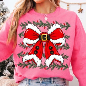 Puede incluir: Una sudadera de cuello redondo en color rosa brillante con un gr&aacute;fico grande de un lazo rojo estilo Santa Claus con borde de piel blanca, un cintur&oacute;n negro con hebilla dorada y cuatro botones negros, enmarcado por ramas de pino y luces rojas.