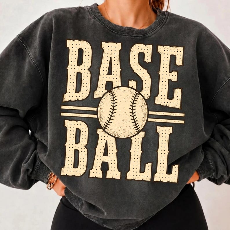 Puede incluir: Una sudadera de cuello redondo en gris carb&oacute;n con efecto lavado mineral, que presenta el texto BASE BALL en una fuente serif grande, punteada y de color crema. Un gr&aacute;fico de una pelota de b&eacute;isbol con costuras visibles est&aacute; centrado.