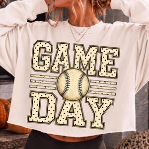 May include: A cream-coloured long-sleeve sweatshirt featuring a graphic print with the text "GAME DAY" in a black-dotted varsity font. A central illustration of a baseball replaces the letter A, with horizontal lines flanking the ball.