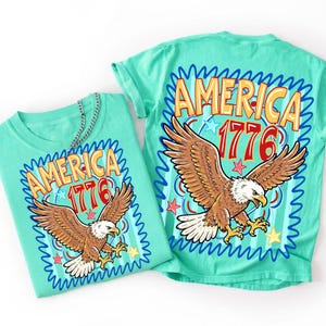 Może przedstawiać: Miętowy t-shirt z nadrukiem graficznym przedstawiającym bielika amerykańskiego w locie oraz napis AMERICA 1776 w kolorze ż&oacute;łtym i czerwonym. Wz&oacute;r zawiera niebieskie faliste obramowania, małe gwiazdki i okrągły motyw. Koszulka jest złożona i płaska.
