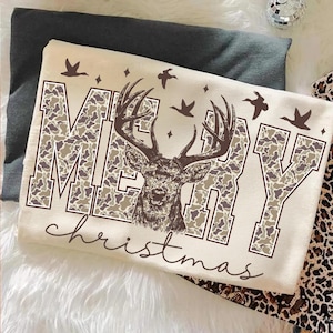 May include: Cream-colored t-shirt with a deer graphic and the words "MERRY Christmas" in a camouflage pattern. The design includes flying birds and stars, creating a festive holiday theme.