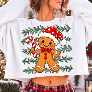 Puede incluir: Una sudadera blanca de manga larga con un gr&aacute;fico central de un hombre de jengibre con un gorro de Pap&aacute; Noel rojo con lunares blancos y una pajarita roja, sosteniendo un bast&oacute;n de caramelo rojo y blanco sobre un fondo de ramas de pino.