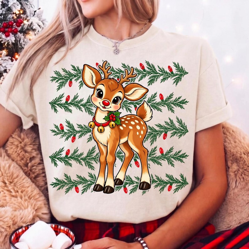 May include: A cream-colored t-shirt featuring a graphic of a brown fawn with large eyes, wearing a collar with gold bells and holly. The background of the graphic consists of horizontal rows of green pine branches interspersed with small red light bulbs.