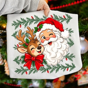 May include: A white t-shirt featuring a graphic illustration of Santa Claus and a reindeer with a large red bow. The design is framed by green pine branches and small red lights, creating a festive holiday scene on the fabric.
