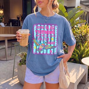 Pu&ograve; includere: Una t-shirt blu a maniche corte con la grafica di un'anatra colorata in volo e il testo "SOME GLAD MORNING WHEN THIS LIFE IS OVER I'LL FLY AWAY" circondato da un bordo rosa e piccole stelle gialle e rosa su uno sfondo a righe azzurre.