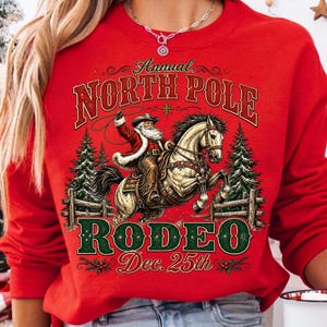 May include: A red long-sleeve sweatshirt featuring a graphic print of Santa Claus riding a horse with a lasso. The text reads Annual NORTH POLE RODEO Dec. 25th. The design includes pine trees, a wooden fence, and decorative star accents.