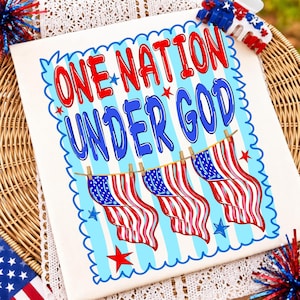 May include: A white t-shirt featuring the red and blue text ONE NATION UNDER GOD. The design includes three American flags hanging from a clothesline with clothespins, surrounded by a blue scalloped border and scattered red and blue stars.