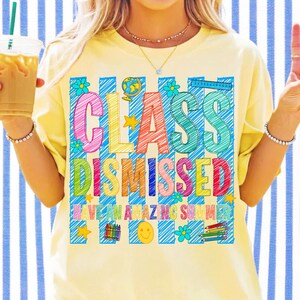 May include: A pale yellow t-shirt featuring a colorful graphic with blue vertical stripes and the text "CLASS DISMISSED" and "HAVE AN AMAZING SUMMER." The design includes illustrations of a globe, flowers, a ruler, crayons, and a smiley face.