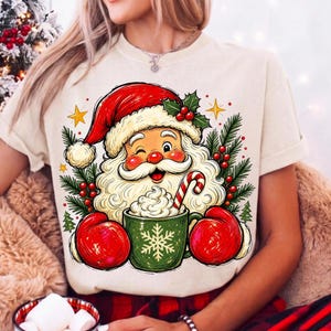 May include: A cream-coloured t-shirt featuring a graphic of Santa Claus wearing a red hat and red mittens, holding a green mug with a white snowflake design and a candy cane. The illustration includes pine branches, red berries, and yellow stars.