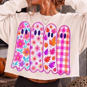May include: A cream-colored long-sleeve sweatshirt featuring a graphic of four ghosts. The first ghost has pink and blue flowers, the second has small pink flowers, the third has orange and purple autumn leaves, and the fourth has a pink and orange plaid pattern.