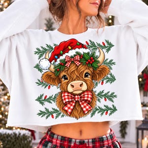 Puede incluir: Una sudadera blanca corta con el gr&aacute;fico de una vaca Highland marr&oacute;n que lleva un gorro de Pap&aacute; Noel rojo con luces de colores, una corona de acebo y un lazo a cuadros rojo y blanco. El fondo incluye ramas de pino verdes y luces rojas.