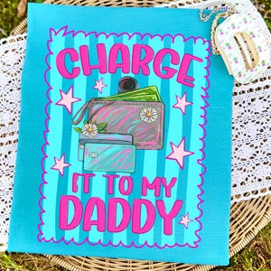May include: A turquoise t-shirt featuring a graphic with vertical stripes and a scalloped border. The design includes a wallet with green paper money, two credit cards, and white daisies. The text reads, "CHARGE IT TO MY DADDY" in pink.