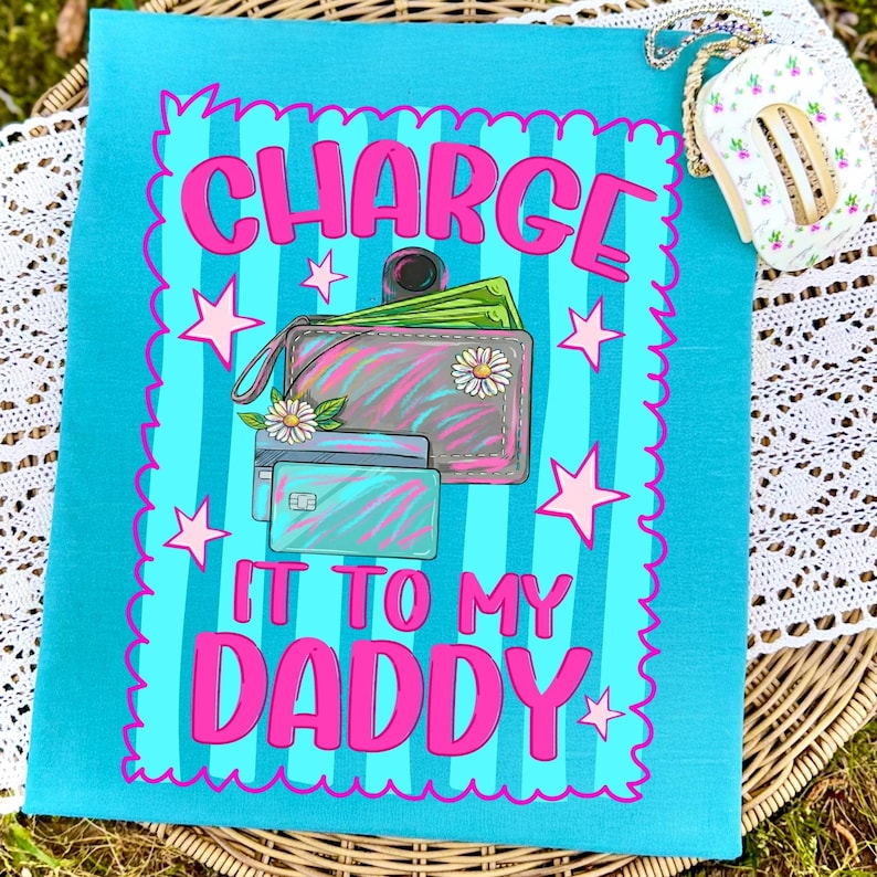 May include: A turquoise t-shirt featuring a graphic with vertical stripes and a scalloped border. The design includes a wallet with green paper money, two credit cards, and white daisies. The text reads, "CHARGE IT TO MY DADDY" in pink.