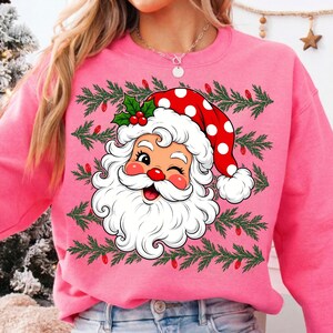 Puede incluir: Una sudadera de cuello redondo en color rosa brillante con un gr&aacute;fico central de un Santa Claus gui&ntilde;ando un ojo, con un gorro rojo de lunares blancos, hojas de acebo verdes y bayas rojas, sobre un fondo de ramas de pino y luces rojas.