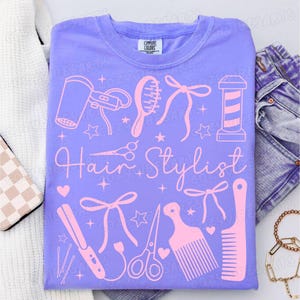 May include: A lilac-colored t-shirt with a pink graphic design featuring hairstyling tools and the words "Hair Stylist". The design includes a hair dryer, comb, scissors, and a barber pole.