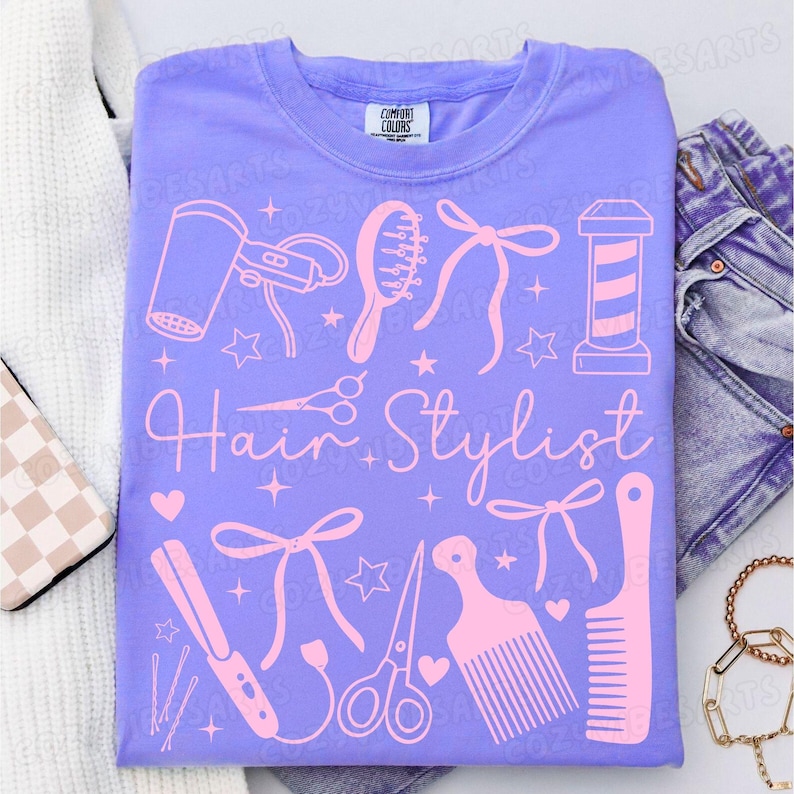 May include: A lilac-colored t-shirt with a pink graphic design featuring hairstyling tools and the words "Hair Stylist". The design includes a hair dryer, comb, scissors, and a barber pole.