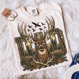 May include: An ivory t-shirt featuring a graphic of a deer head with large antlers centred over the word DAD in block letters. The design includes a forest landscape, flying birds, and pine branches. A tag is attached to the collar area.