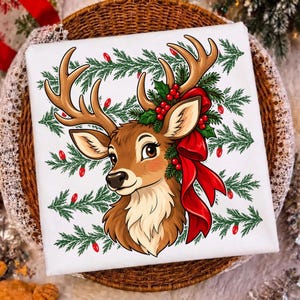May include: A white t-shirt featuring a graphic illustration of a brown deer with large antlers, adorned with a bright red bow, green pine branches, and red berries, set against a background pattern of green pine needles and small red lights.