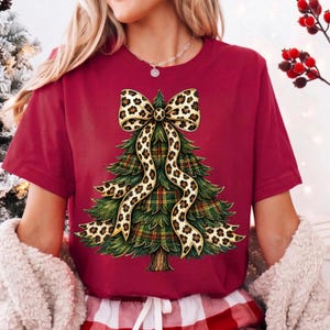 May include: A maroon short-sleeved t-shirt featuring a graphic of a green Christmas tree with a plaid pattern. The tree is adorned with a large, flowing ribbon featuring a tan and black leopard print pattern tied in a bow at the top.