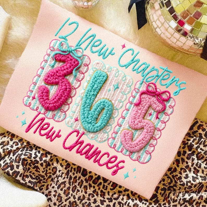 Puede incluir: Un coj&iacute;n rectangular rosa con texto y n&uacute;meros bordados. El texto dice "12 New Chapters 365 New Chances" y los n&uacute;meros son "365". Los n&uacute;meros son rosas y turquesas, con un borde decorativo.