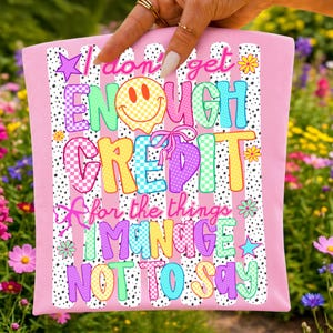 Pode incluir: Um artigo de tecido rosa claro com um fundo de riscas de bolinhas pretas e brancas e texto colorido que diz I don't get ENOUGH CREDIT for the things I MANAGE NOT TO SAY, acentuado com uma cara sorridente amarela e pequenas flores.