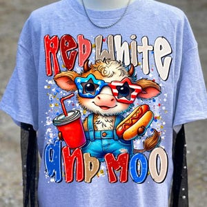 Puede incluir: Una camiseta gris claro con el gr&aacute;fico de una vaca de dibujos animados que lleva gafas de sol en forma de estrella con patrones rojos, blancos y azules. La vaca sostiene una bebida roja y un perrito caliente. El texto dice RED WHITE AND MOO.