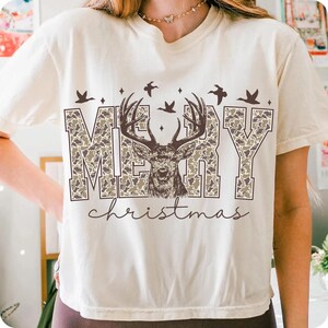 May include: Cream-colored t-shirt with a "Merry Christmas" design. The text is in a camouflage pattern, with a deer head in the center. Above the text are flying birds, creating a festive holiday theme.