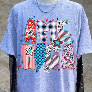 May include: A light grey t-shirt featuring the word AMERICA in large, multi-patterned letters. The design includes red, white, and blue colors with star, checkerboard, striped, and spotted patterns, accented by various small star shapes.