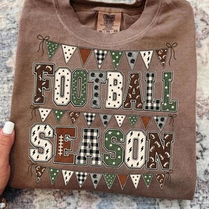 May include: A brown crew neck t-shirt featuring the text FOOTBALL SEASON in large block letters. The letters are filled with various patterns including stars, buffalo plaid, and footballs. Bunting banners with similar patterns frame the text.
