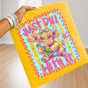 Puede incluir: Una manta amarilla con una ilustraci&oacute;n colorida de una vaca highland sosteniendo un frasco de pepinillos. El texto "JUST DILL WITH IT" est&aacute; impreso en letras rosas alrededor de la vaca. El dise&ntilde;o incluye un borde festoneado rosa y estrellas.