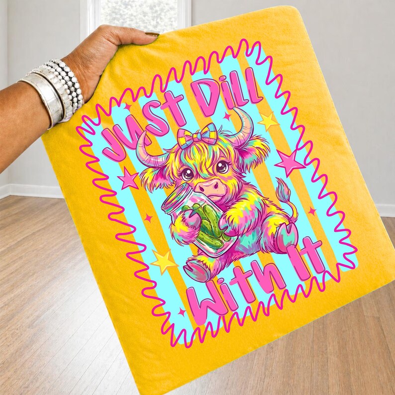 Puede incluir: Una manta amarilla con una ilustraci&oacute;n colorida de una vaca highland sosteniendo un frasco de pepinillos. El texto "JUST DILL WITH IT" est&aacute; impreso en letras rosas alrededor de la vaca. El dise&ntilde;o incluye un borde festoneado rosa y estrellas.