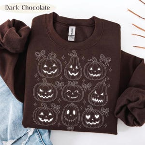 May include: A dark chocolate brown crewneck sweatshirt featuring a white line art graphic of various jack-o'-lanterns, pumpkins, and small bows. The design includes multiple pumpkin shapes with different carved facial expressions and star accents.