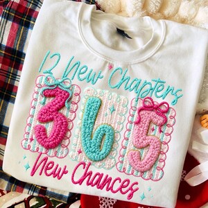 Puede incluir: Sudadera blanca con texto y n&uacute;meros bordados. El dise&ntilde;o incluye las frases "12 New Chapters" y "365 New Chances" en turquesa y rosa. Los n&uacute;meros 3, 6 y 5 son de estilo crochet, con lazos rosas.