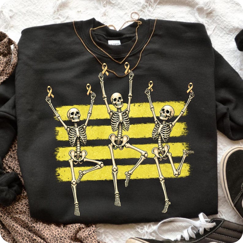 May include: A black crewneck sweatshirt featuring a graphic of three white skeletons dancing in front of three horizontal yellow brushstroke stripes. Each skeleton holds a small yellow awareness ribbon above its head in a celebratory pose.