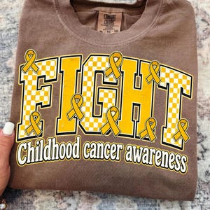 May include: A brown t-shirt featuring the word FIGHT in large yellow and white checkered block letters, accented with multiple yellow awareness ribbons. Below the main text, the words Childhood cancer awareness are printed in white.
