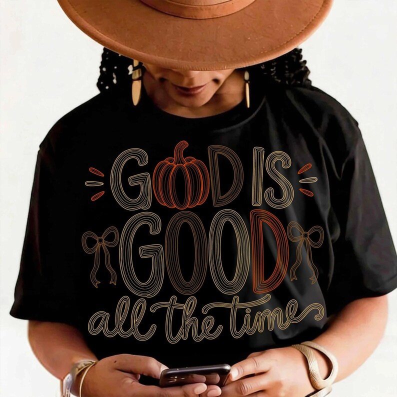 May include: A black t-shirt featuring the text "GOD IS GOOD all the time" in a stylized, multi-line font. The design includes an orange pumpkin graphic replacing the letter O in GOD and two small line-drawn bows on the sides of the word GOOD.