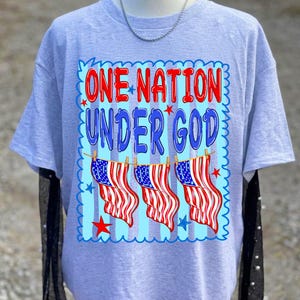 May include: A light grey short-sleeved t-shirt featuring a graphic print with the text ONE NATION UNDER GOD in red and blue letters. The design includes three hanging American flags, small red and blue stars, and a scalloped blue border.