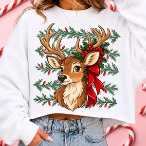 May include: A white cropped sweatshirt featuring a graphic illustration of a brown deer head with large antlers, adorned with green pine branches, red berries, and a large red bow, set against a background of horizontal green pine boughs and red lights.