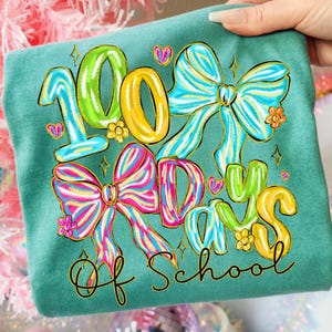 Op de afbeelding: Tealkleurig T-shirt met een kleurrijke afbeelding ter viering van 100 schooldagen. Het ontwerp toont de cijfers "100" in groen en geel, strikken, harten en de woorden "Days of School" in een grillige lettertype.