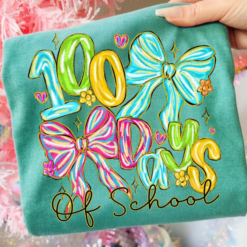 Op de afbeelding: Tealkleurig T-shirt met een kleurrijke afbeelding ter viering van 100 schooldagen. Het ontwerp toont de cijfers "100" in groen en geel, strikken, harten en de woorden "Days of School" in een grillige lettertype.