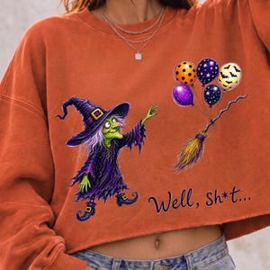 K&ouml;nnte beinhalten: Ein orangefarbenes Langarm-Sweatshirt mit einer Grafik einer gr&uuml;nh&auml;utigen Hexe in violettem Gewand und Hut, die einen Besen mit bunten gemusterten Luftballons h&auml;lt. Der Text "Well, Sh*t..." ist in schwarzer Schrift unter der Grafik gedruckt.
