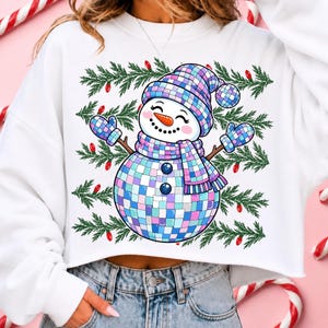 May include: A white long-sleeve sweatshirt featuring a graphic of a snowman made of iridescent disco ball tiles. The snowman wears a matching checkered hat, scarf, and mittens, set against a background of green pine branches and red light bulbs.